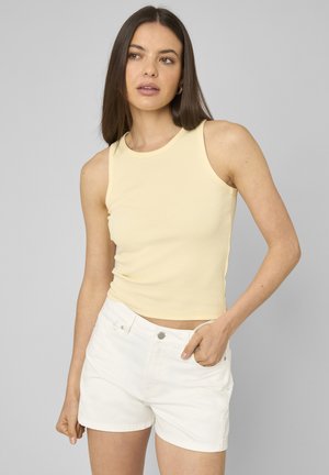 Femme aux longs cheveux bruns portant un haut sans manches jaune pâle et un short blanc, debout devant un fond gris neutre.