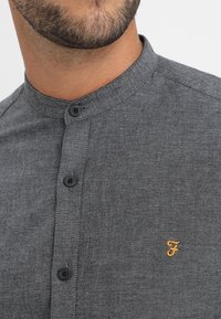 Chemise grise à boutons avec col mandarin, dotée de deux boutons noirs et d'un petit logo doré brodé sur le côté gauche.