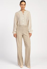 Crème blouse met knoopsluiting en een textuur aan de voorkant, met lange mouwen, gecombineerd met hooggetailleerde beige broek en bijpassende puntige schoenen.