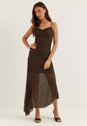 Femme portant une robe en dentelle marron foncé sans manches avec un ourlet asymétrique, associée à des chaussures noires à talons ouverts et des bijoux en or.