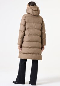 Abrigo tipo acolchado marrón con capucha, que presenta un diseño de acolchado y un dobladillo hasta la rodilla. Textura suave y acolchada con mangas anchas. Pantalones negros.