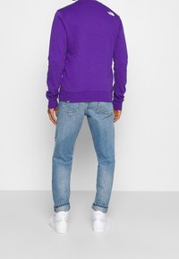 Fialová mikina s žebrovanými manžetami a lemem, v kombinaci se světle modrými džínami. Na horní části zad výrazné logo, nošena s bílými teniskami.