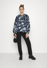 Blusa azul marinha com tie-dye, mangas balão, decote em forma de fenda, calças pretas e sapatos pretos robustos. Pequena bolsa preta texturizada.