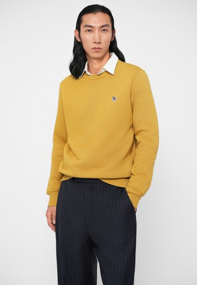 Κίτρινη μπλούζα με στρογγυλή λαιμόκοψη και κεντητό λογότυπο, συνδυασμένη με σκούρα παντελόνια με ρίγες. Λείος υφάνσεις, άνετη εφαρμογή.