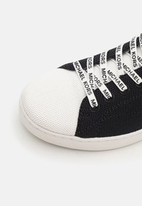 Lähikuva musta-valgetest spordijalatsitest, millel on "Michael Kors" kaubamärgiga valged paelad valgel tallal, ühetoonilise tausta ees.
