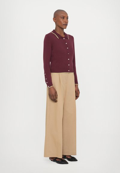 Cardigan bordeaux avec un col et des bordures jaunes contrastantes, doté de boutons sur le devant. Associé à un pantalon beige à jambes larges et des chaussures noires.