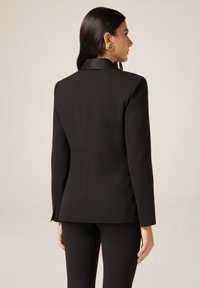 Czarna marynarka o dopasowanym kroju, z ostrymi klapami i dopasowanymi szwami. Gładka tkanina z długimi rękawami, w zestawieniu z czarnymi spodniami.