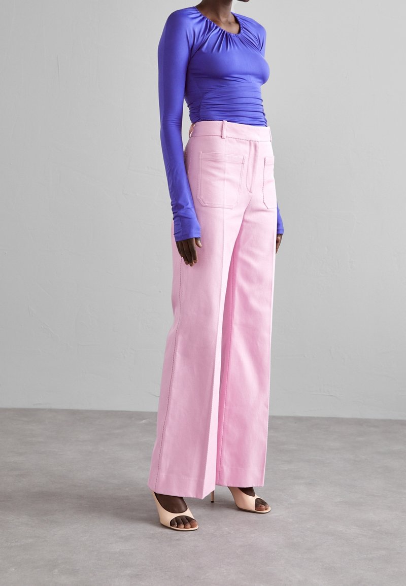 Fialová blůza s dlouhým rukávem a nařasením, v kombinaci s výrazně růžovými kalhotami s vysokým pasem a širokými nohavicemi, doplněná béžovými sandály na podpatku s otevřenou špičkou. Viditelné textury látky.