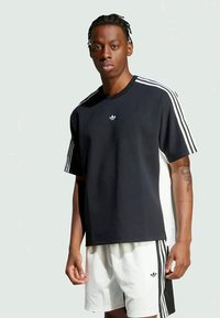 Jauns vīrietis ar pītas matiem, ģērbies melnā Adidas kreklā ar baltām svītrām un baltās Adidas šortos ar melnām svītrām, stāv pret vienkāršu fonu.