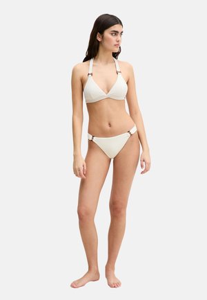 Kobieta stojąca boso, ubrana w kremowy, teksturowany strój bikini z detalami w formie pierścieni na ramiączkach i majtkach, patrząca w prawo na jednolitym tle.