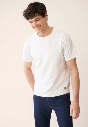 Homme souriant aux cheveux foncés portant un t-shirt blanc uni à manches courtes et un jean bleu foncé, debout devant un fond beige clair.