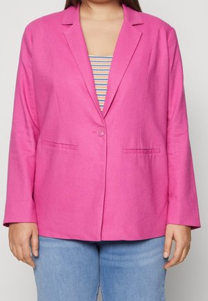 Person iført en knallpink enkeltknappet blazer over en stribet top og lyseblå jeans, vist fra brystet til hofterne.