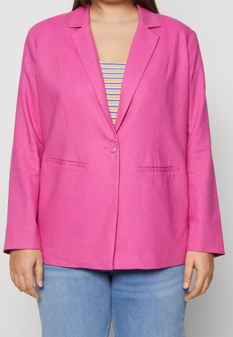 Personne portant un blazer rose vif à un bouton sur un haut rayé et un jean bleu clair, montré de la poitrine aux hanches.