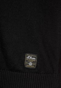 Maglione nero lavorato a maglia con tessuto strutturato e una targhetta rettangolare del marchio s.Oliver in giallo e nero nella parte inferiore.