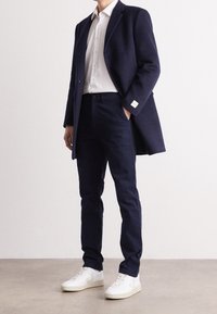 Man med marinblå kostym, vit skjorta och vita sneakers, stående på en enkel golv mot en vit vägg.