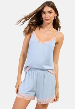 Woman wearing light blue sleeveless top and shorts set with pale pink lace trim, standing against plain white background.