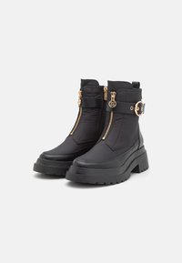 Botas de tobillo negras hechas de textil y caucho, con cremalleras doradas, una suela ancha y gruesa, y una correa con hebilla que tiene un acento redondo.