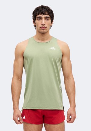 Man die een lichtgroen mouwloos sportshirt en rode hardloopshorts draagt, staand met een neutrale gezichtsuitdrukking tegen een effen achtergrond.