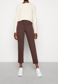 Pantaloni in cotone marrone con vestibilità sartoriale, cuciture a contrasto e chiusura con bottone. Abbinati a un maglione color crema e sneakers bianche.