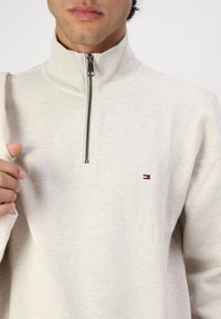 Lichtbeige sweatvest met rits en hoge kraag, gemaakt van zacht materiaal. Bevat een klein geborduurd logo in rood, wit en blauw.