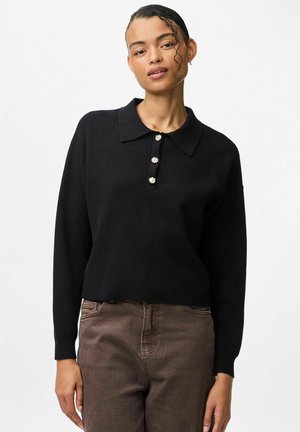 Un pull noir avec un col, des manches longues et quatre boutons ronds en métal à l'avant. Le tissu semble tricoté et doux. Associé à un pantalon marron.