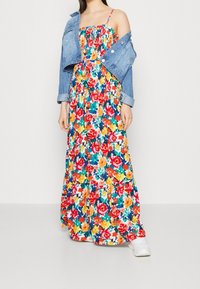 Robe longue à motif floral comprenant du rouge, jaune et vert. Assortie à une veste en jean, dévoilant des bretelles et un corsage froncé.