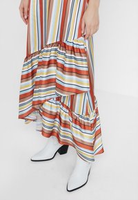 Kobieta w wielokolorowej, pasiastym spódnicy z falbanami, nosząca białe, szpiczaste botki za kostkę, stojąca na jednolitym białym tle.