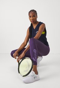 Marineærmeløs topp, lilla leggings med "Lacoste"-logo, hvite joggesko med lyseblå detaljer, holder en grønn og svart tennisracket.