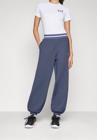 Hvit kortermet t-skjorte med svart EA7-logo, kombinert med løstsittende navy joggebukser med stripete midjebånd og sidelommer.