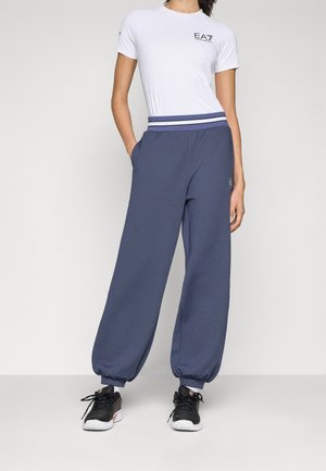T-shirt bianca a maniche corte con logo EA7 nero, abbinata a joggers blu navy a vestibilità ampia con cintura a righe e tasche laterali.