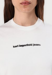 White cotton t-shirt featuring a blurred black logo “karl lagerfeld jeans” across the chest, with a ribbed crew neckline and short sleeves.