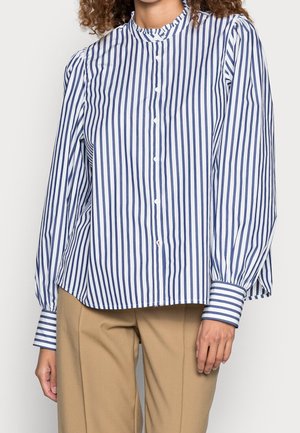 Kobieta ubrana w białą koszulę zapinaną na guziki z pionowymi granatowymi paskami i beżowe spodnie, stojąca na tle jednolitego, jasnego tła.