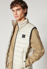 Helle beigen gepolsterten Weste mit hohem Kragen, ausgestattet mit einem schwarzen Logo-Patch, Reißverschluss und Seitentaschen; getragen über einem braunen Langarmshirt.