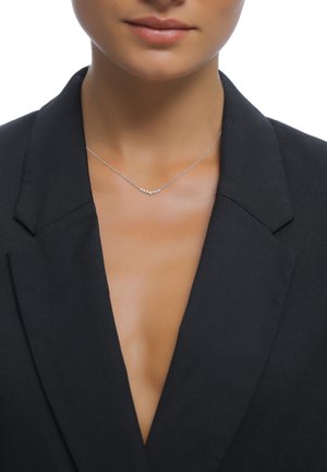 Sølvhalskæde med et buet design, der har fem små ædelsten, båret med en sort blazer og en dyb udskæring. Glat metallovertag.