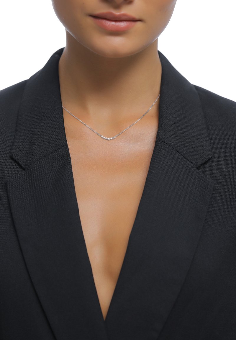 Sølvhalskæde med et buet design, der har fem små ædelsten, båret med en sort blazer og en dyb udskæring. Glat metallovertag.