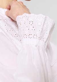 Weiße Bluse mit hochgeschlossenem Spitzenkragen und puffigen bestickten Ärmeln. Der Stoff ist leicht und strukturiert mit floralen Mustern.