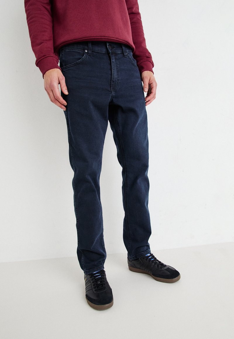 Jeans din denim închis la culoare, cu croială slim, având cinci buzunare, detalii subtile de cusătură și fiind purtați cu pantofi sport negri.