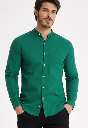 Homme portant une chemise verte ajustée à col mao avec des boutons blancs, un pantalon noir, les mains dans les poches, debout devant un fond blanc.