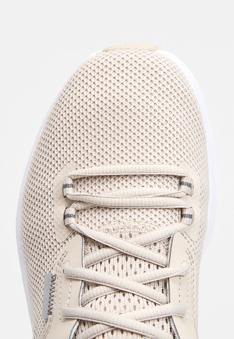 Zapatilla deportiva de color beige claro con parte superior de malla texturizada, cordones elásticos y una suela blanca suave. Diseño minimalista con acentos sutiles.