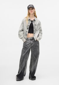 Jasnoszara denimowa kurtka z kieszenią na piersi, czarny krótki top i czarne szerokie dżinsy z wyblakłym wzorem, do tego ciemna czapka.