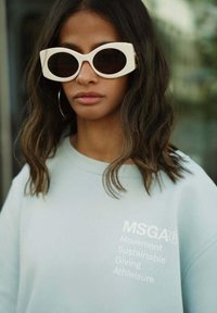 Lichtblauwe oversized sweatshirt met witte tekst "MSGA® Movement Sustainable Giving Athleisure" en grote witte zonnebril.