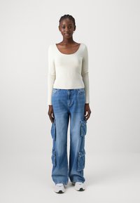 Hosszú ujjú krém színű blúz lekerekített nyakkivágással, amelyet kék, széles szárú cargo farmernadrággal párosítanak, több zsebbel és elhalványult denim hatással.