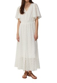 Donna che indossa un maxi vestito bianco a trama con maniche a farfalla e vita arricciata, abbinato a sandali piatti con cinturino alla caviglia marroni.