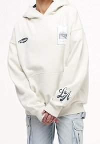 Ljust beige hoodie i mjukt tyg, med en framficka, ett grafiskt tryck på bröstet och "LA"-detalj på ärmen.