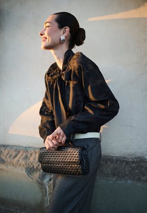 Une femme souriante, vêtue d’un chemisier noir à motifs et d’un pantalon gris, tient un sac à main noir tissé devant un mur de pierre ensoleillé.