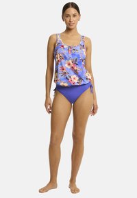 Een model staat vol vertrouwen in een bloemige tankini-top en een blauwe bikinionderbroek, waarmee een levendige zwemmode stijl wordt getoond tegen een eenvoudige achtergrond.