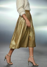 Goldene plissierte Midi-Rock mit glänzender Textur, kombiniert mit metallischen Absatzsandalen. Der Pullover hat eine flauschige, helle Farbe.