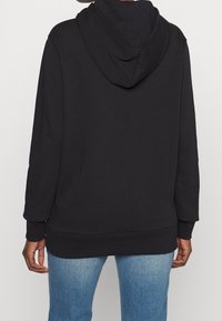 Zwarte capuchontrui met een losse pasvorm, zachte textuur en lange mouwen. Voorzien van een capuchon met trekkoord en geribbelde manchetten en zoom.
