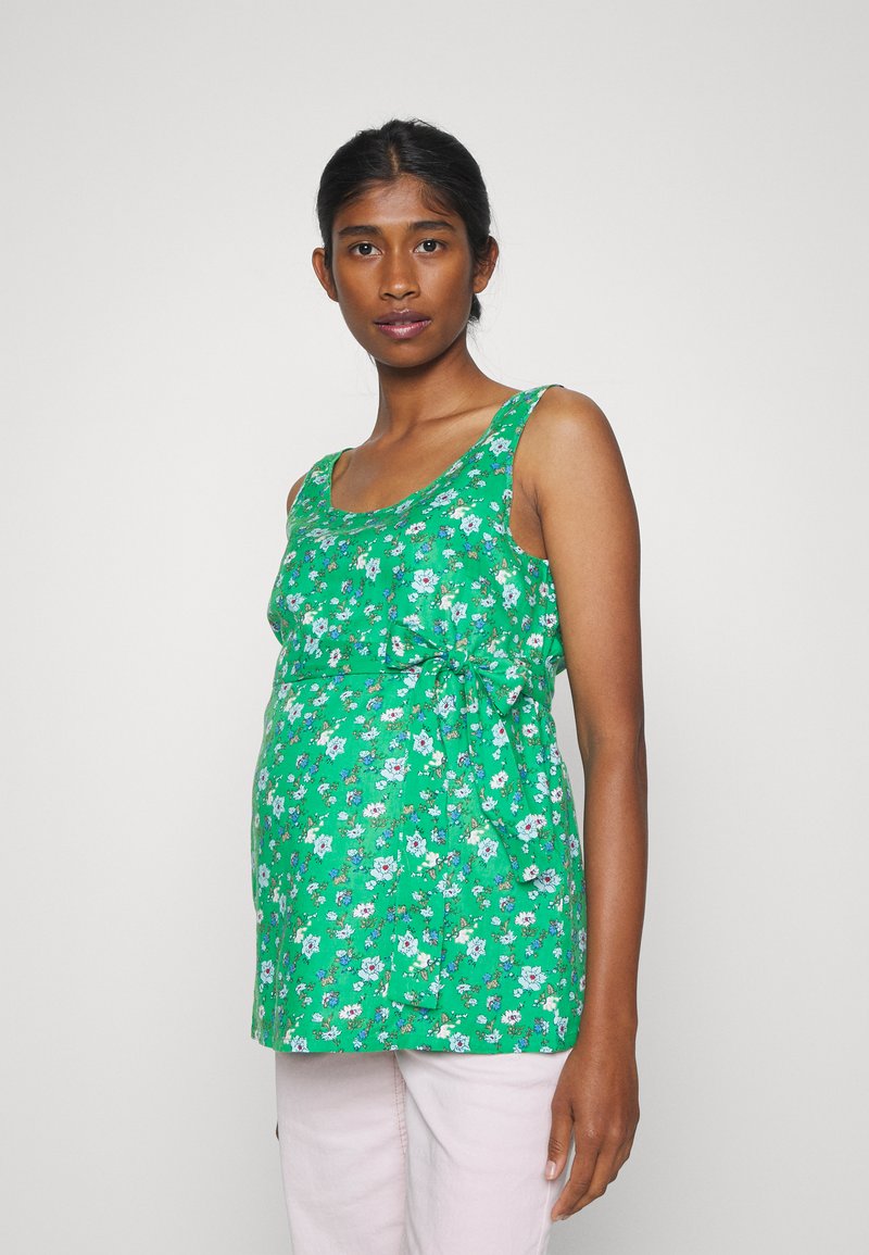 Blusa de maternidade verde floral com um corte solto, decote arredondado e detalhe em laço lateral. Apresenta pequenas flores brancas e azuis por toda a peça.