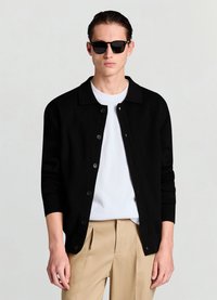 Cardigan en maille noire avec un col chemise, fermeture par boutons, et poignets côtelés, porté sur une chemise blanche, associé à un pantalon beige.